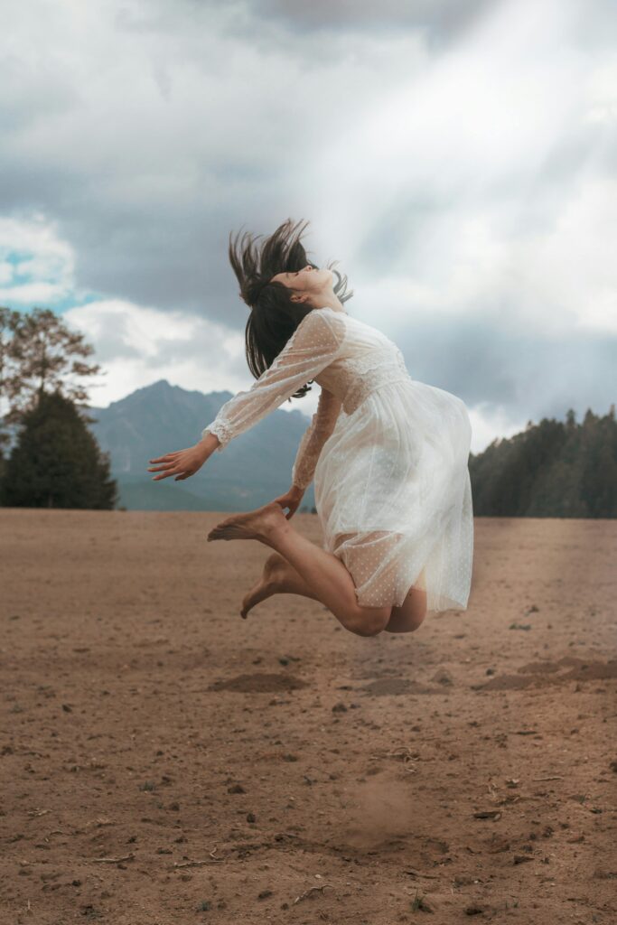 A woman in a flowing white dress jumps joyfully in a picturesque mountain landscape under a cloudy sky.
